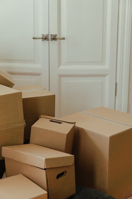 A collection of cardboard boxes of various sizes stacked and placed in front of a white double door with metal handles, inside a room. The boxes are made of plain brown corrugated cardboard, some with