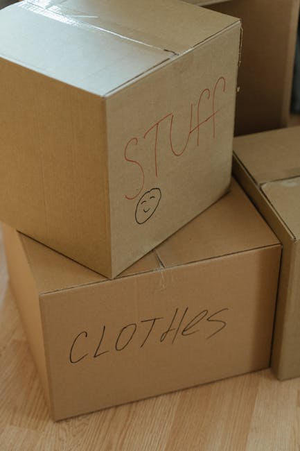 A collection of cardboard boxes of various sizes stacked and placed in front of a white double door with metal handles, inside a room. The boxes are made of plain brown corrugated cardboard, some with