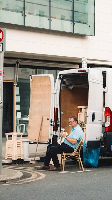 A man wearing glasses and a light blue shirt is seated on a wooden chair beside a white delivery van parked on the street outside a modern building. The van's rear doors are open, revealing interior s
